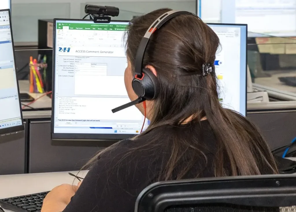 Rear view of a woman wearing a headset at a desk, looking at a computer monitor with a form open in a busy office/call center.