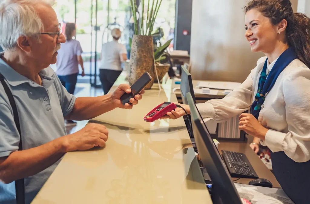 Older man holding a smartphone while a receptionist offers a card reader across a counter at a hotel front desk.