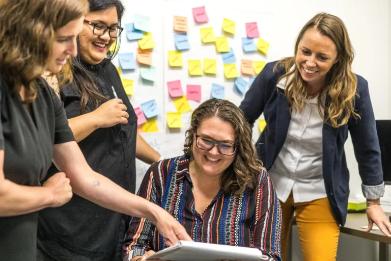Four coworkers smile and review a tablet together in an office, with colorful sticky notes covering a whiteboard in the background.